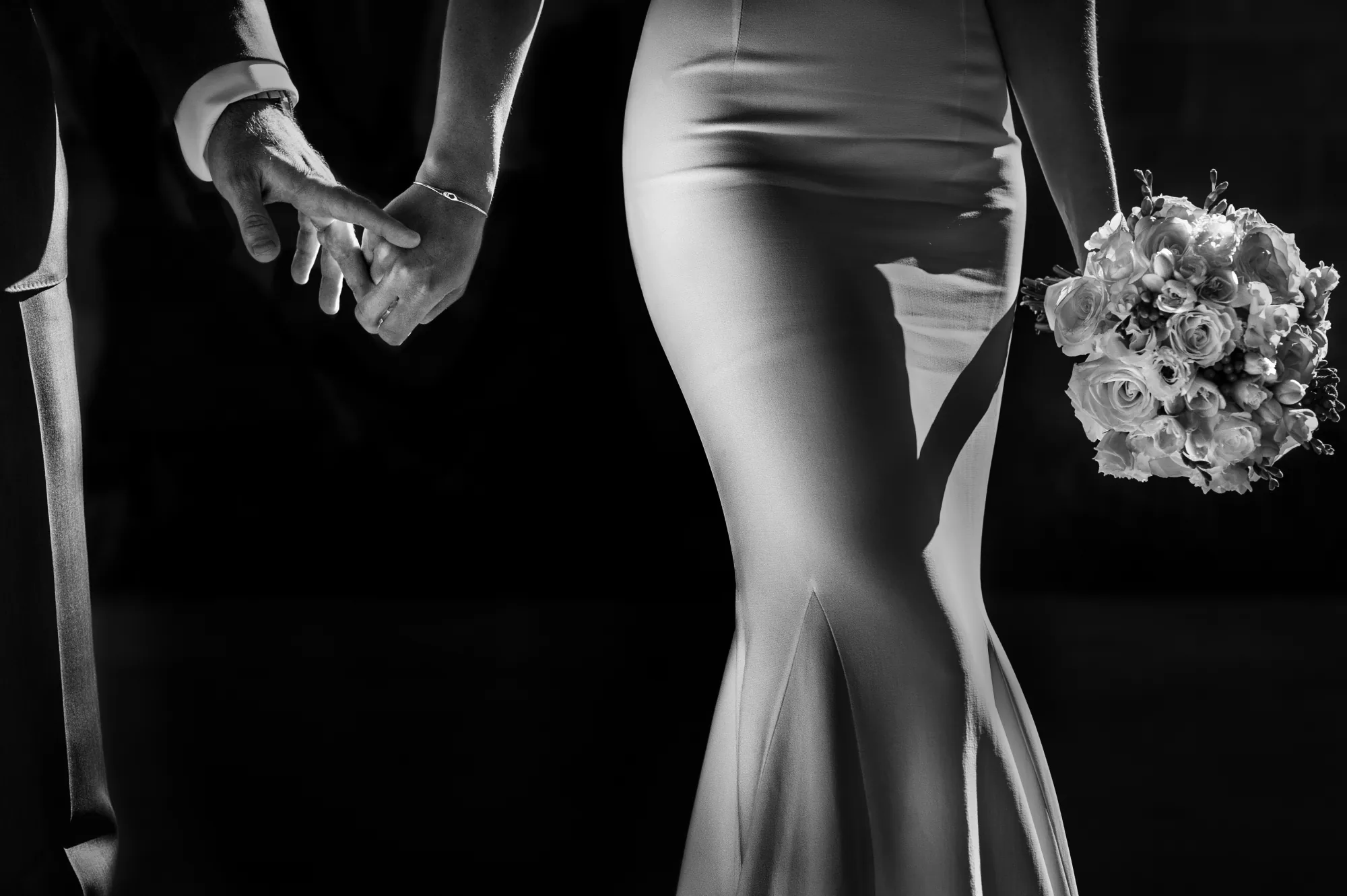 Black and white detail of a bride in a satin gown, holding hands and her bouquet