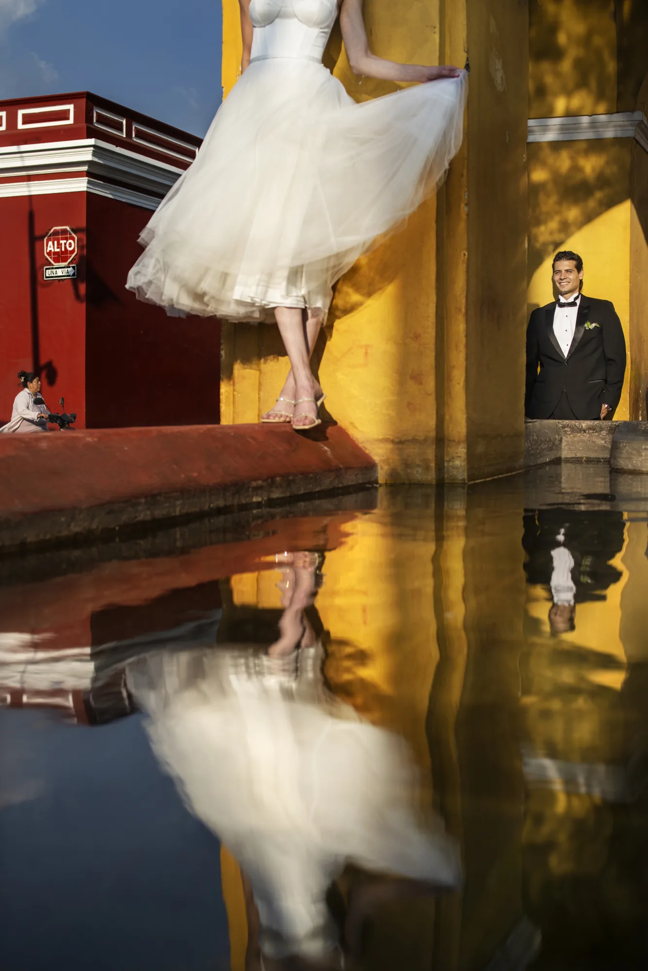 Bride and groom reflected in water before a yellow and red building