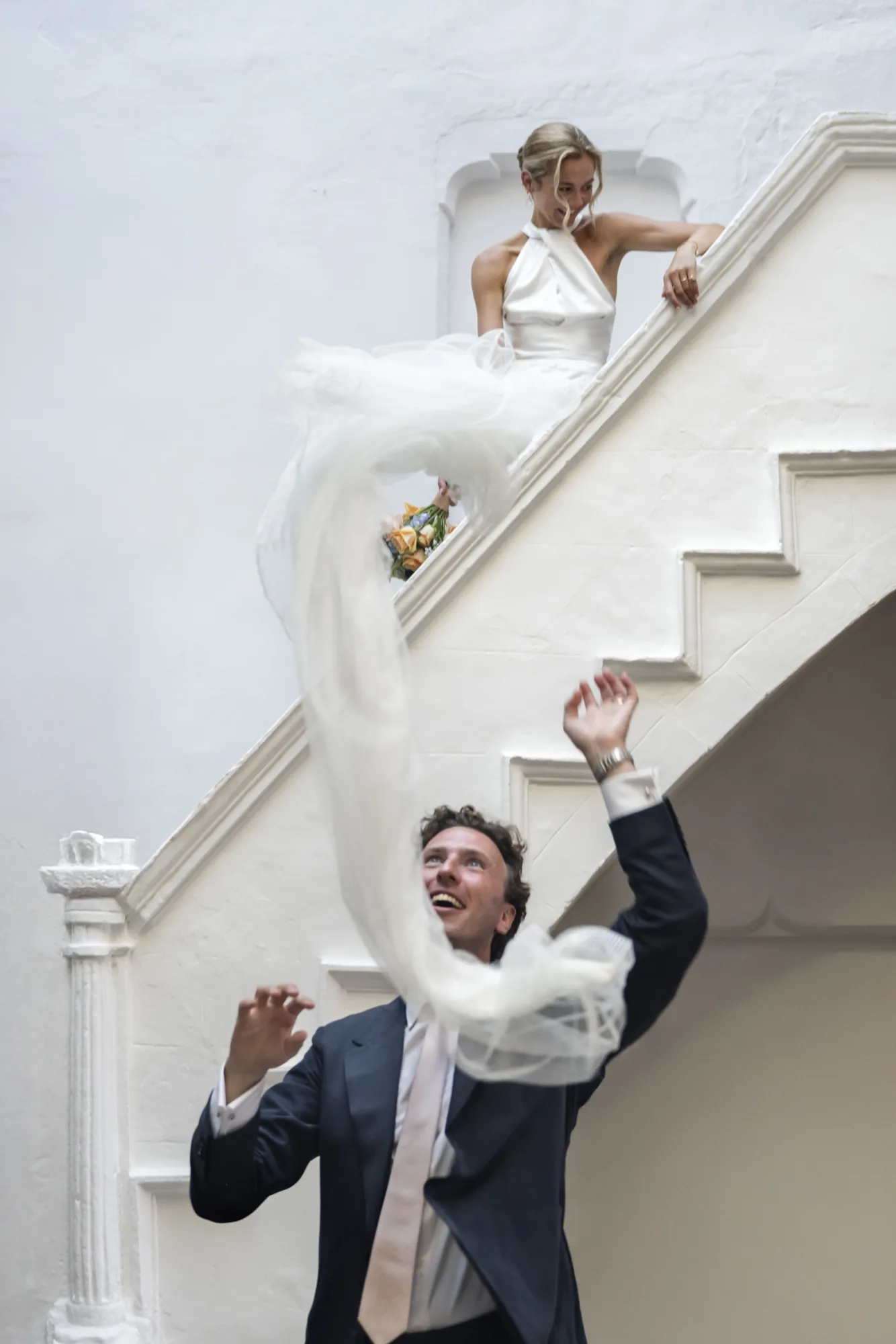 Bride on white stairs as the groom reaches up to her