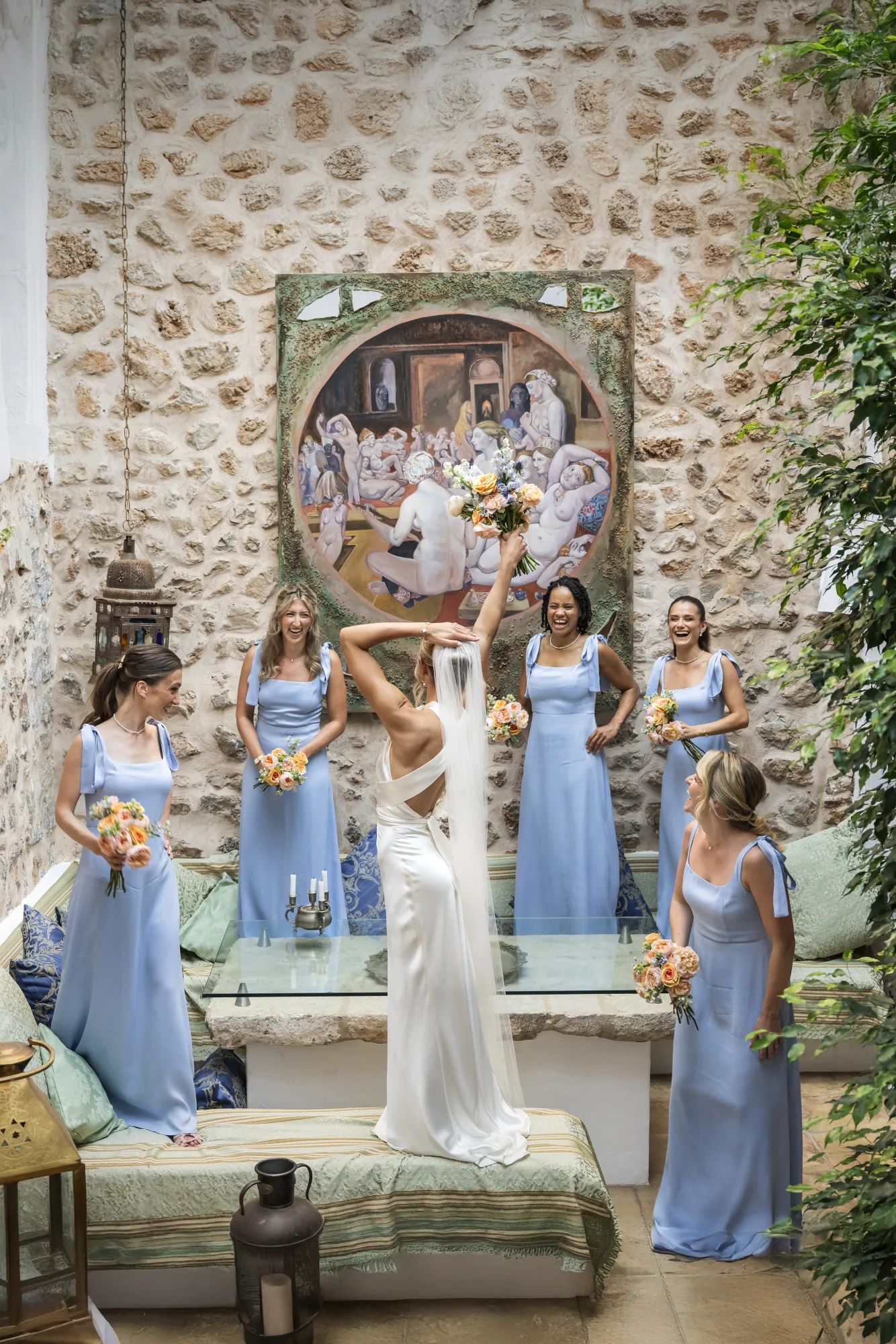 Bride raising her arms among bridesmaids in pale blue in a stone courtyard