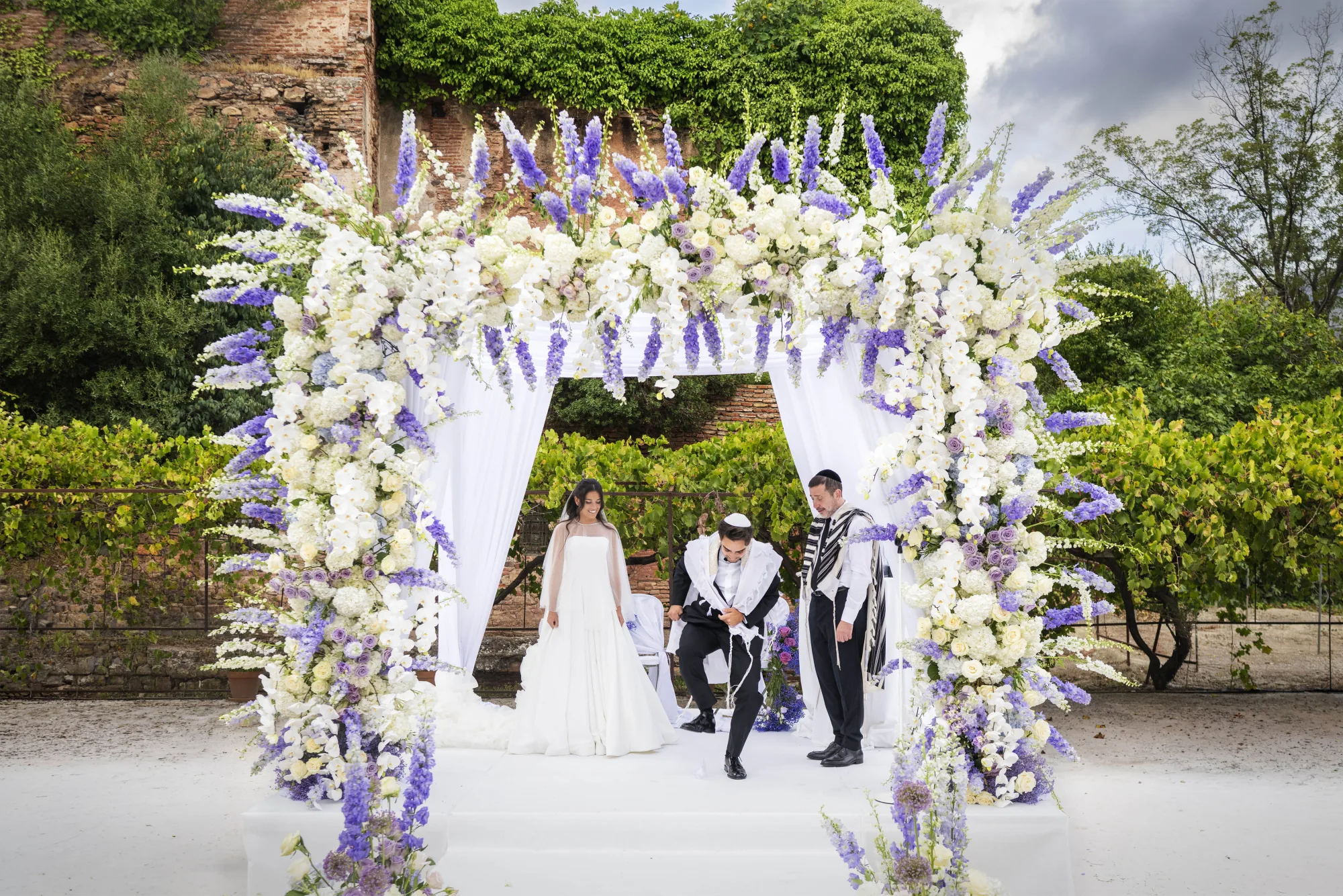 Couple under a white and lilac floral arch at a garden ceremony