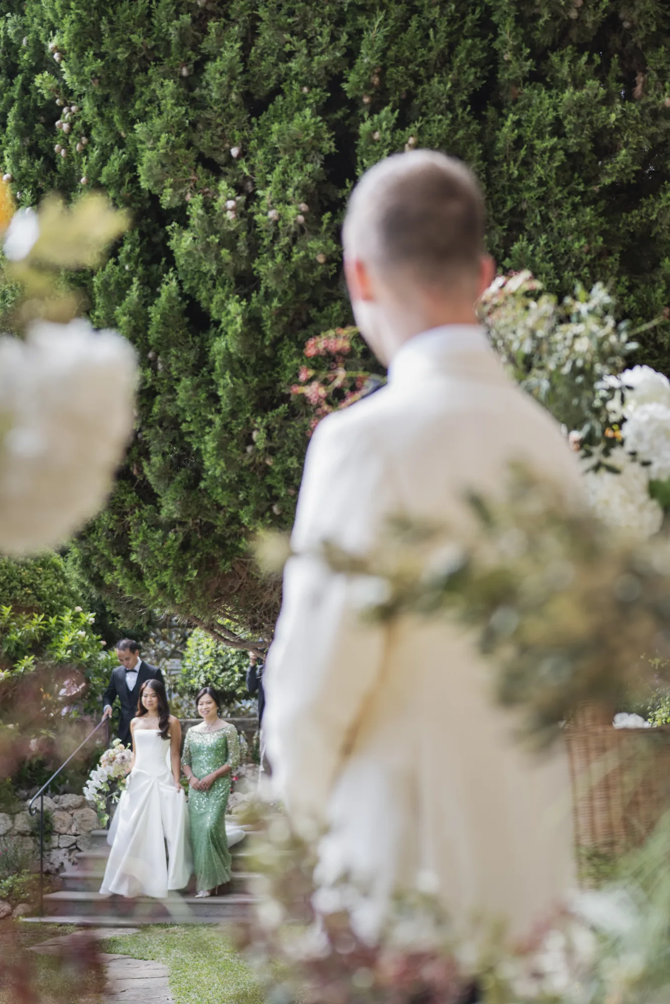 Groom in a white jacket watching the bride arrive through the greenery