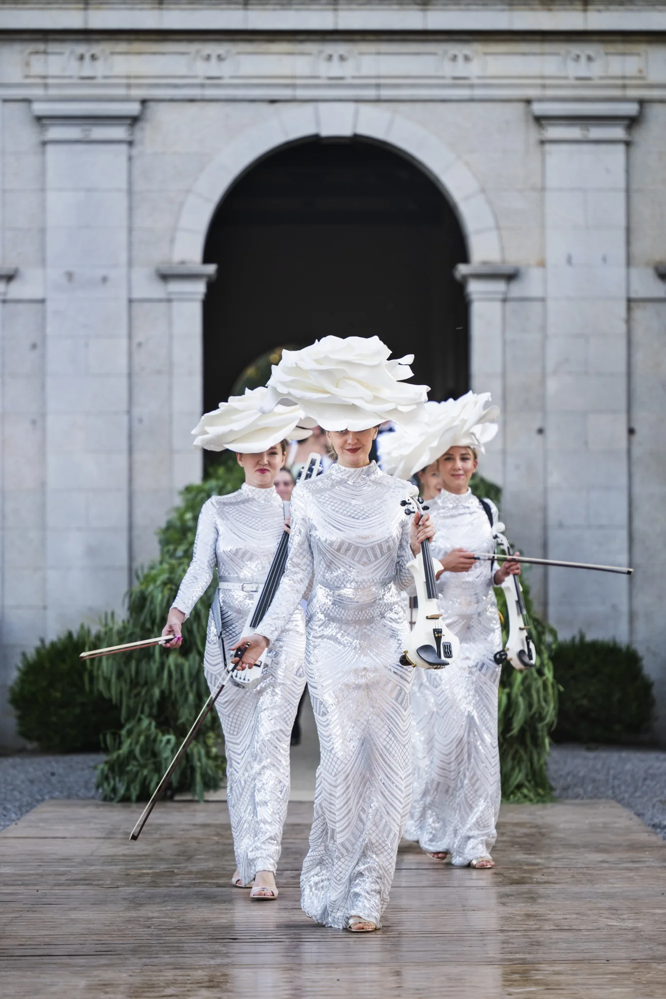 Guests in white suits and giant white flower hats walking out of a stone archway