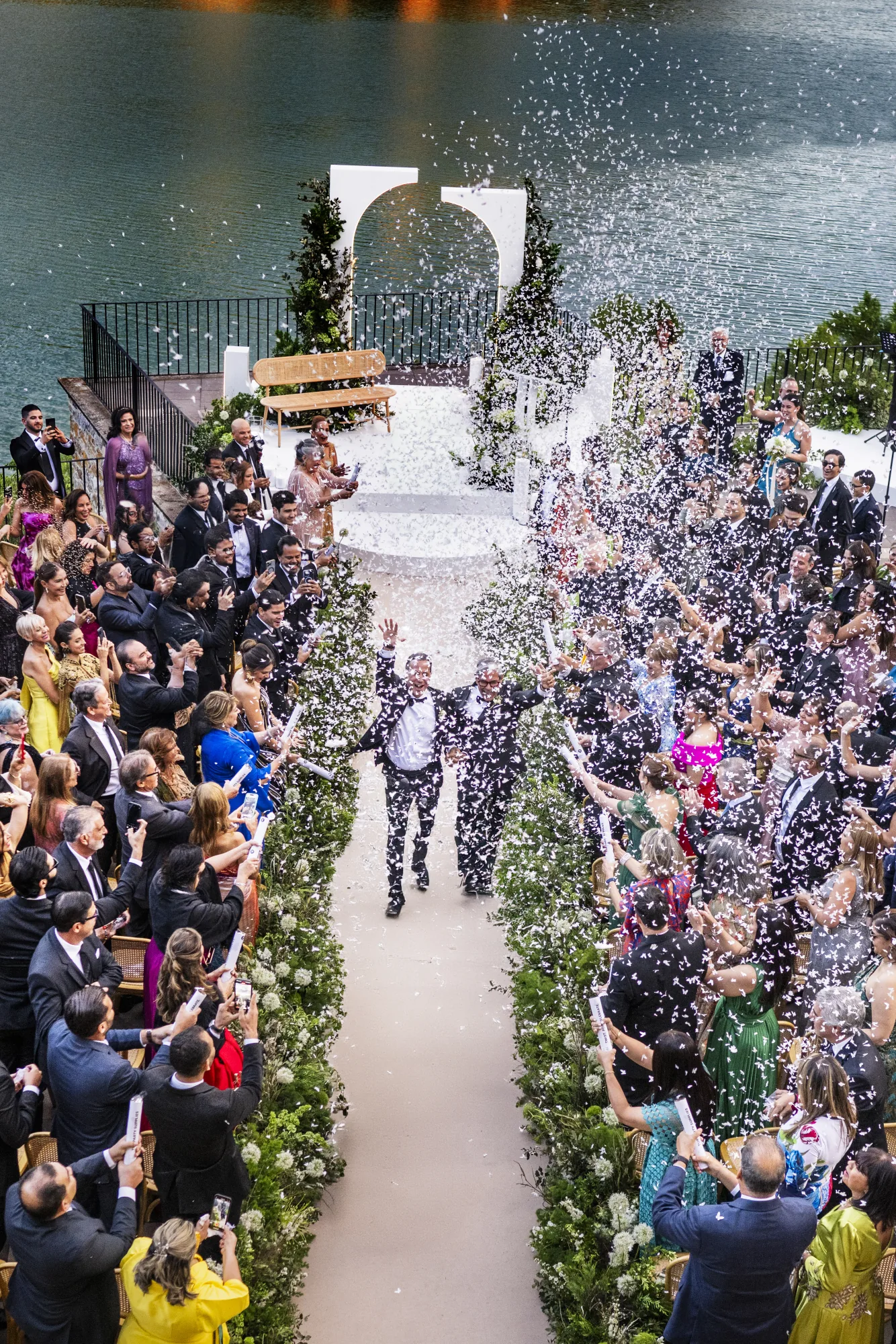 Guests throwing confetti over the couple along a lakeside aisle