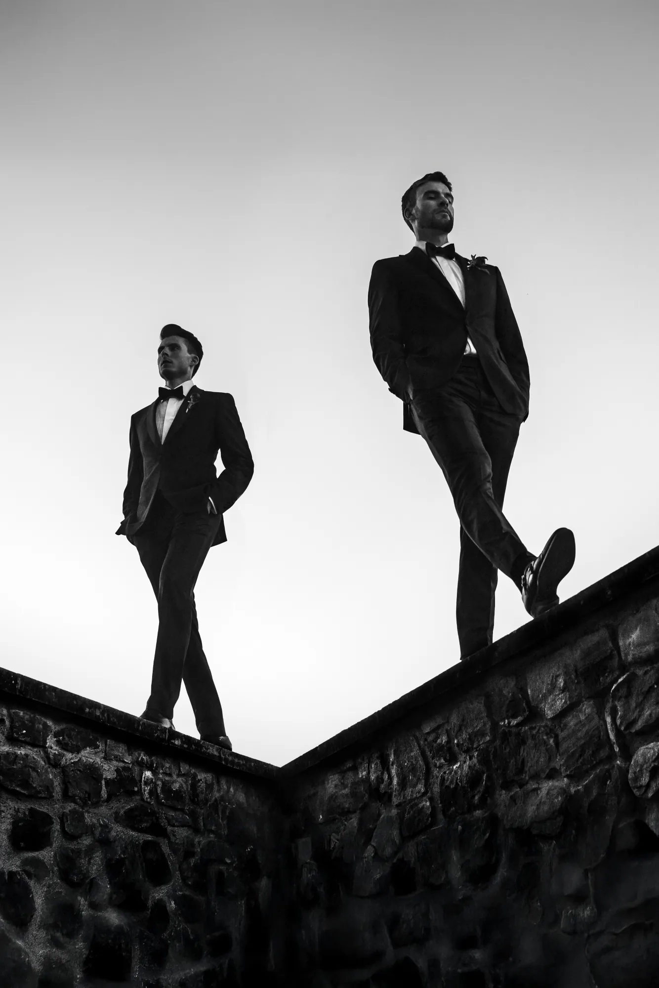 Two grooms in black tie walking along a stone wall against a pale sky