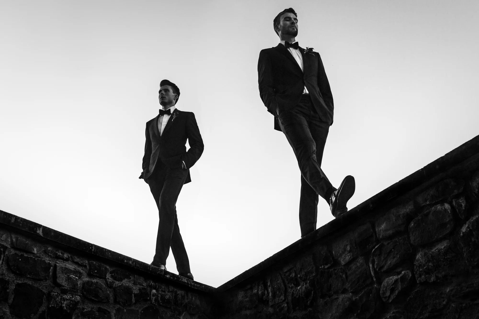 Two grooms in tuxedos silhouetted against a bright sky on a stone staircase