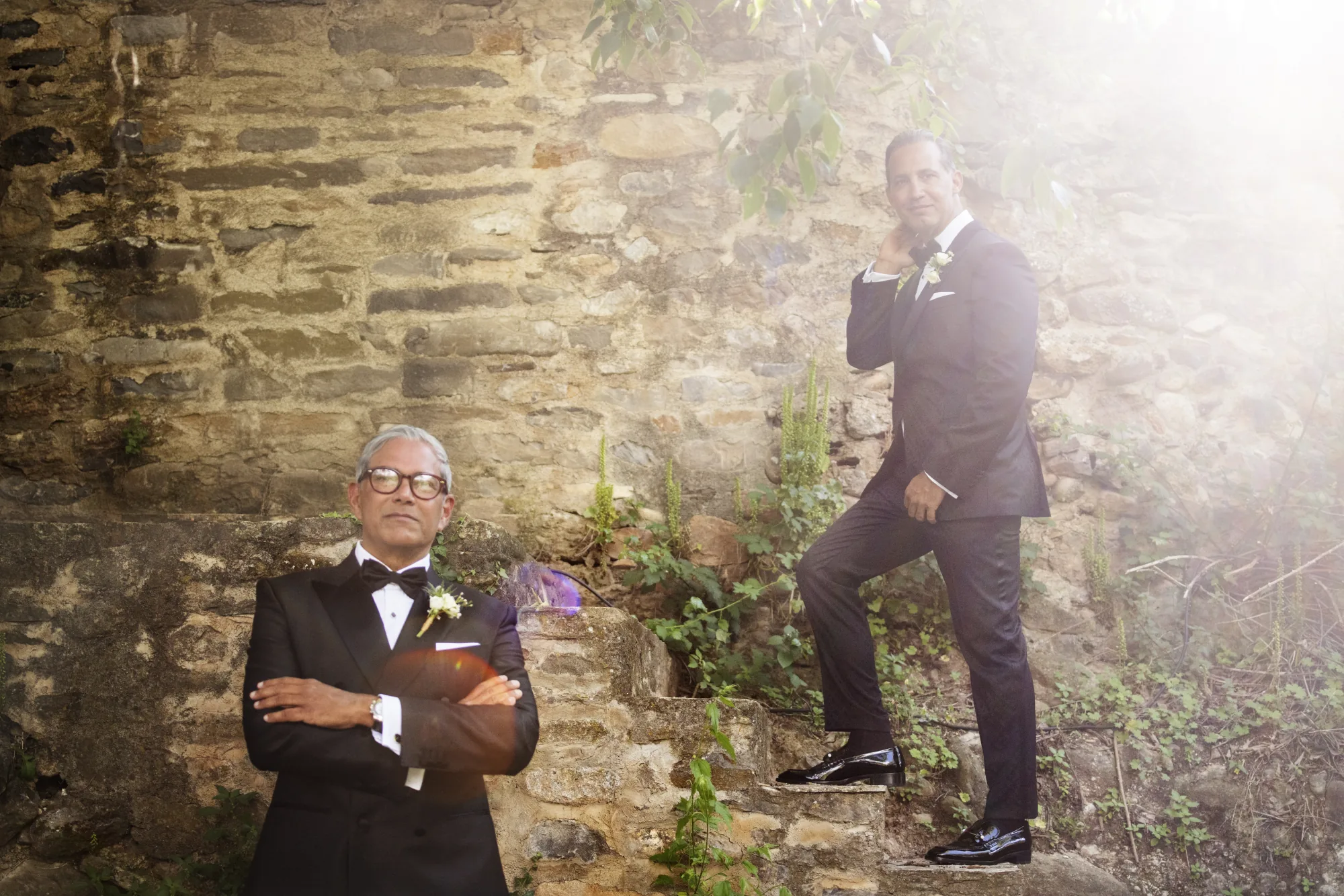 Two men in black tie posing against a sunlit stone wall